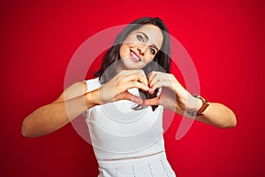 Young beautiful woman wearing white dress standing over red isolated background smiling in love doing heart symbol shape with