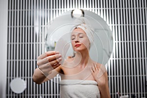 Young beautiful woman wearing white towel holding pink gouache scraper for face massage while doing beauty morning routine in