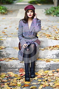 Young beautiful woman wearing winter coat and cap sitting on steps full of autumn leaves