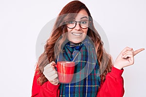 Young beautiful woman wearing winter scarf and drinking a cup of hot coffee smiling happy pointing with hand and finger to the