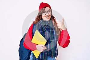 Young beautiful woman wearing winter scarf and student backpack holding book smiling happy and positive, thumb up doing excellent