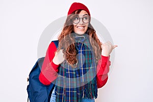 Young beautiful woman wearing winter scarf and student backpack pointing thumb up to the side smiling happy with open mouth
