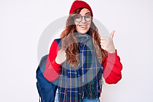 Young beautiful woman wearing winter scarf and student backpack smiling happy and positive, thumb up doing excellent and approval