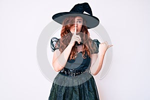 Young beautiful woman wearing witch halloween costume asking to be quiet with finger on lips pointing with hand to the side
