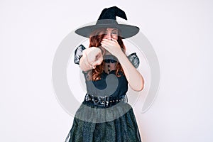 Young beautiful woman wearing witch halloween costume laughing at you, pointing finger to the camera with hand over mouth, shame