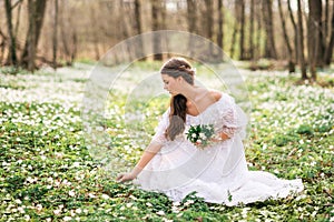 Young beautiful woman in a white dress collects primroses. A girl in the spring forest. A bouquet of white anemones in her hands