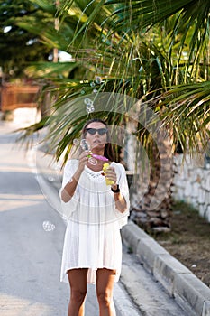 Young beautiful woman in white dress and sunglasses blowing soap bubbles on the road with palms. The concept of joy