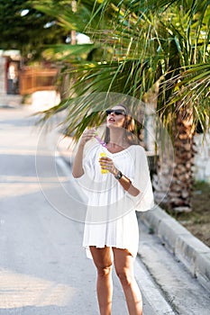 Young beautiful woman in white dress and sunglasses blowing soap bubbles on the road with palms. The concept of joy