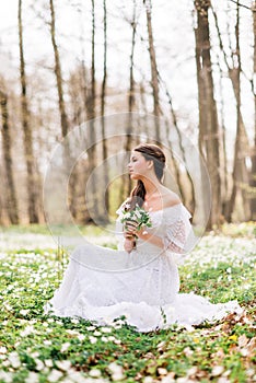 Young beautiful woman in a white long dress collects primroses. Fairy girl in the spring forest. A bouquet of white flowers in her