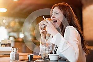 Young beautiful woman in white stylish blouse looks hungry eating meat burger with fries for lunch in trendy cafe eating outside