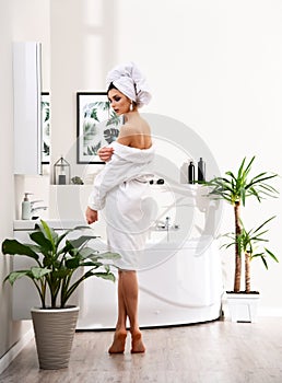 Young beautiful woman with white towel on head standing near bathtub wearing bathrobe in modern bathroom