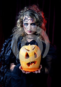 Young beautiful woman in a witch costume with bright makeup and hairstyle holding a pumpkin, a symbol of halloween.