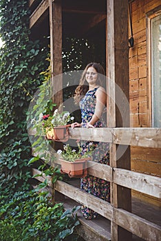 Young beautiful woman on a wooden balcony, holding hands on the railing. Around her nature and flowers.