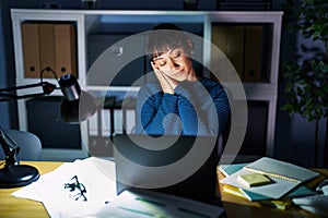 Young beautiful woman working at the office at night sleeping tired dreaming and posing with hands together while smiling with