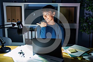 Young beautiful woman working at the office at night smiling with happy face looking and pointing to the side with thumb up