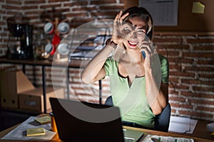 Young beautiful woman working at the office at night speaking on the phone doing ok gesture with hand smiling, eye looking through