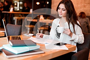 Young beautiful woman working outside office in her laptop taking notes in her planner while drinking hot coffee in the cafe