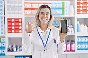 Young beautiful woman working at pharmacy drugstore showing smartphone screen smiling happy and positive, thumb up doing excellent