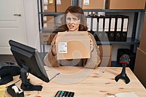 Young beautiful woman working at small business ecommerce holding box clueless and confused expression