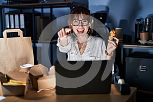 Young beautiful woman working using computer laptop and eating delivery food pointing to you and the camera with fingers, smiling