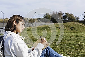 Young beautiful woman writing in notebook outdoors in public park, taking notes