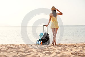 Young beautiful woman in yellow dress and hat with large suitcase on tropical beach. Girl look on sea