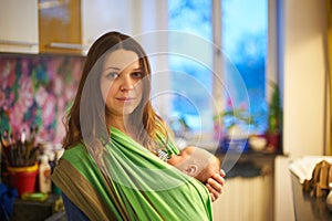 Young woman with newborn infant baby in a sling with a blurred background at home