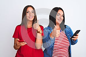 Young beautiful women using smartphone standing over isolated white background happy with big smile doing ok sign, thumb up with