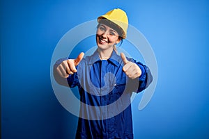 Young beautiful worker woman with blue eyes wearing security helmet and uniform approving doing positive gesture with hand, thumbs