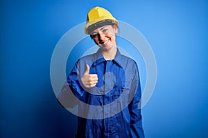 Young beautiful worker woman with blue eyes wearing security helmet and uniform doing happy thumbs up gesture with hand