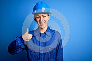 Young beautiful worker woman with blue eyes wearing security helmet and uniform doing happy thumbs up gesture with hand
