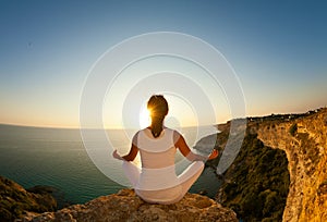 Young beautiful yoga woman meditating in lotus pose on the precipice of a cliff overlooking the sea.