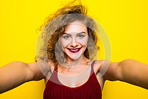 Young beautifulgirl with an curly hairstyle. Laughing girl take selfie from phone on Yellow background.