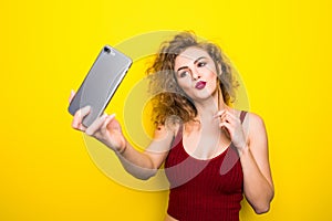 Young beautifulgirl with an curly hairstyle send selfie kiss. Laughing girl take selfie from phone on Yellow background.