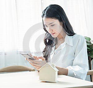 Young beauty Asian business woman in casual hand using smart mobile phone sit at home with house model, planing to buy or rent