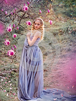 Young beauty blonde in a flying lilac dress in the garden where there are many flowering trees with pink magnolia. Copy space.