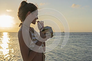 Young beauty girl drinking coconut at tropical beach near sea water at paradise island at sunset. Summer concept. Holiday travel