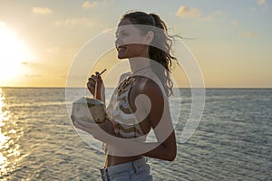 Young beauty girl drinking coconut at tropical beach near sea water at paradise island at sunset. Summer concept. Holiday travel