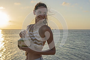 Young beauty girl drinking coconut at tropical beach near sea water at paradise island at sunset. Summer concept. Holiday travel