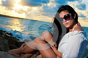 Young beauty with sunglasses sitting on a rock by the ocean