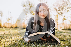 Young believing girl lying on the grass and studying her bible, in the field at sunset. Copy space