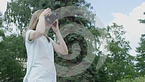 Young bespectacled blonde woman takes pictures in park on summer day leaning forward and backward looking for the best