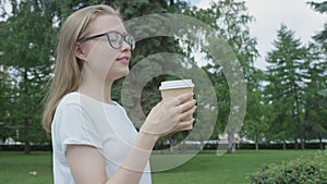 Young bespectacled young woman drinking a cup of coffee in park on summer day calmly and confidently looking away from