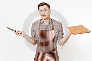 Young bewildered man chef or waiter in striped brown apron, shirt holding wooden cutting board, knife isolated on white