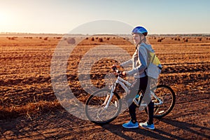 Young bicyclist riding in autumn field with haystacks at sunset. Woman with backpack travelling on bike