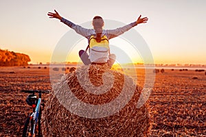 Young bicyclist sitting on haystack with raised and opened arms after a ride. Woman having rest in autumn field.