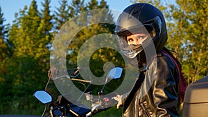 Young biker girl riding touring motorbike on forest road. Beautiful woman with red hair in leather jacket sitting on motorcycle in