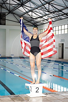 Young biracial female swimmer holding American flag, standing by pool indoors