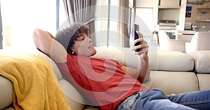 A young biracial man lounges on a sofa at home, smartphone in hand
