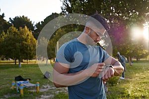 Young biracial man in t-shirt looking down at fitness tracker by trees in park